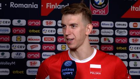 Artur Szalpuk w czerwonej koszulce reprezentacji Polski udziela wywiadu na tle ścianki sponsor&oacute;w.