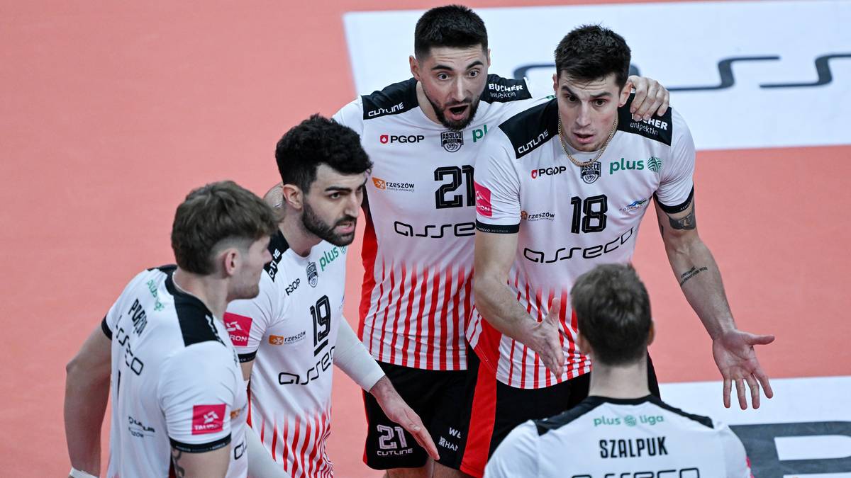 Grupa siatkarzy w białych strojach z czerwonymi pasami, z numerami na koszulkach, dyskutuje na boisku.