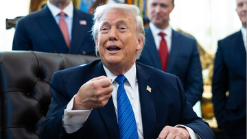 Donald Trump siedzi przy biurku, m&oacute;wiąc i gestykulując.