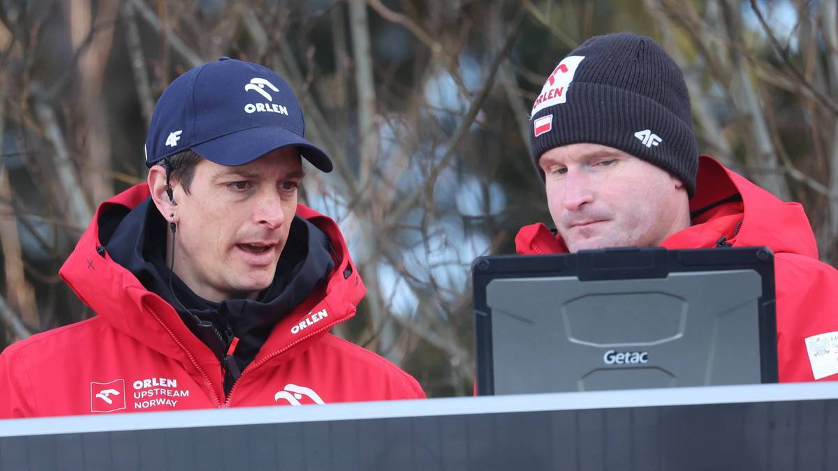 Dwóch mężczyzn w czerwonych kurtkach i czapkach z logo Orlen, jeden z nich patrzy na ekran tabletu.