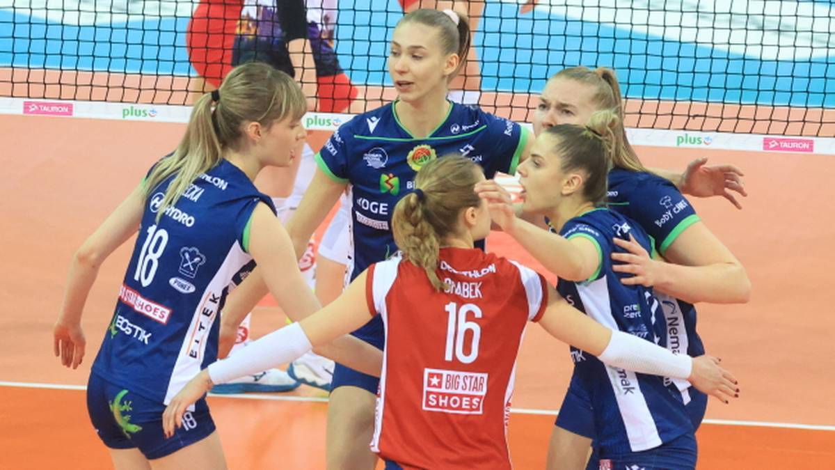 Pięć zawodniczek drużyny siatkarskiej w granatowo-białych strojach otacza zawodniczkę w czerwonym stroju, okrzykując się i dotykając.