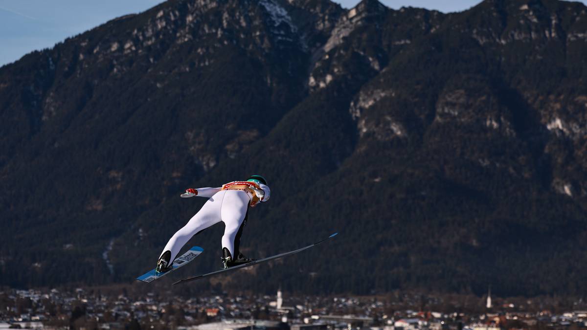 Skoczek narciarski w białym kombinezonie wykonuje długi skok na tle ośnieżonych g&oacute;r i miasta.