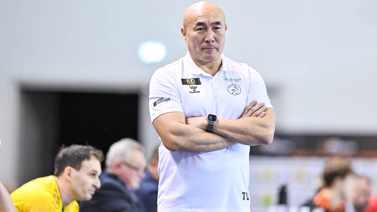 Trener piłki ręcznej w białej koszulce z krótkim rękawem, spogląda przed siebie ze skrzyżowanymi rękami.