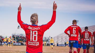 Piłkarz w czerwonej koszulce z numerem 18 na plecach, klęczy na boisku z rękami uniesionymi w g&oacute;rę w geście radości.