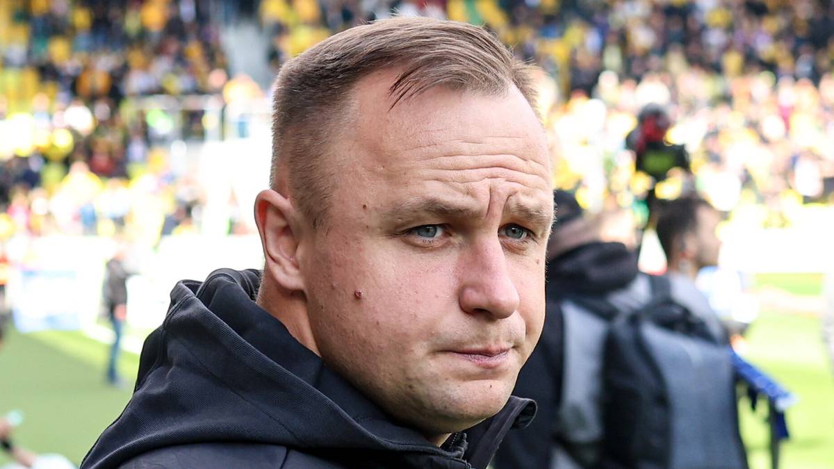 Portret mężczyzny w średnim wieku z krótkimi, jasnymi włosami, patrzącego w bok z lekko zmarszczonymi brwiami. Jest ubrany w ciemną bluzę z kapturem. W tle widać nieostre sylwetki ludzi w strojach sportowych oraz trybuny stadionu z kibicami.