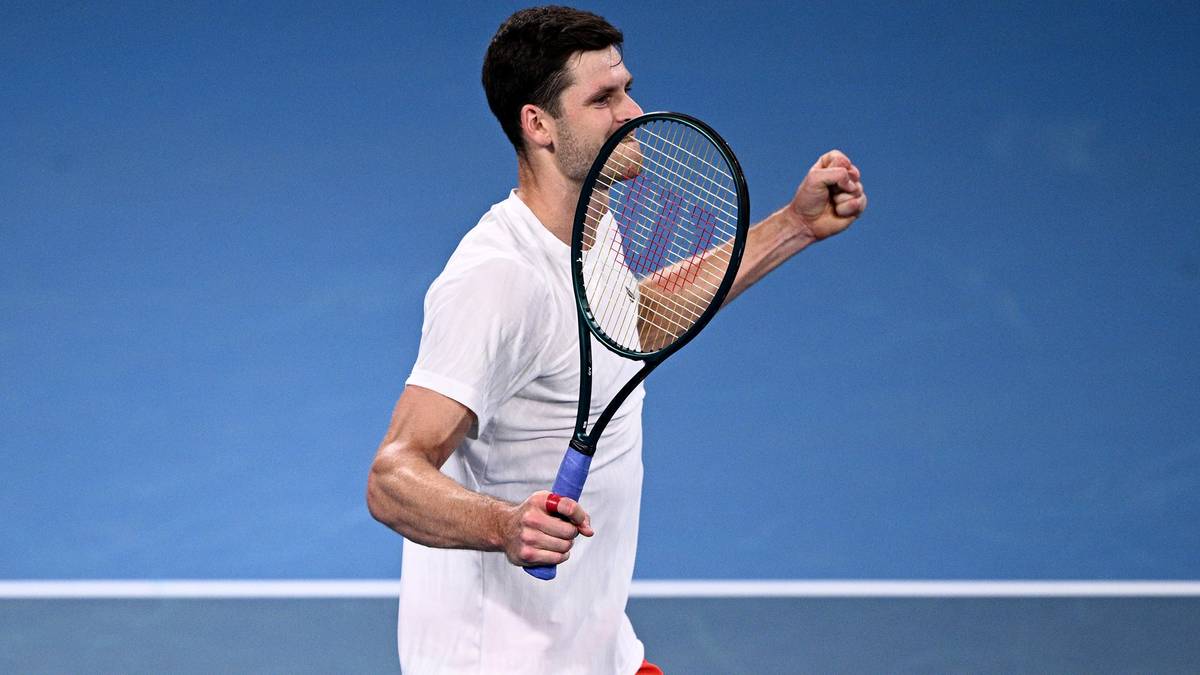 Tenisista w białej koszulce i czerwonych spodenkach, trzymający rakietę i zaciskający pięść.