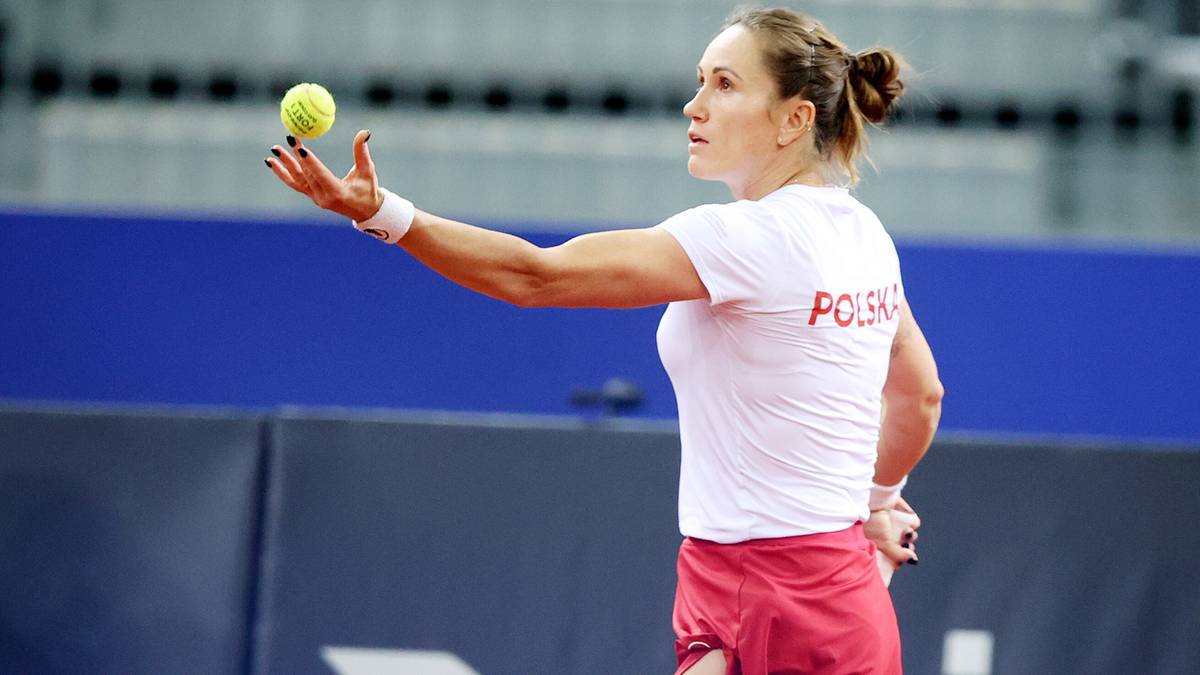 Tenisistka w białej koszulce z napisem "POLSKA" i czerwonej spódniczce serwuje piłkę do tenisa.