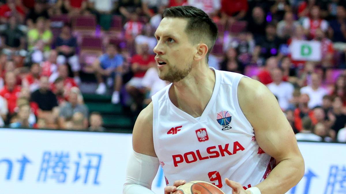 Zawodnik koszykówki w białej koszulce z napisem "POLSKA" i numerem 9, trzymający piłkę, na tle trybun pełnych widzów.