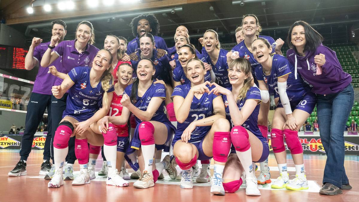 Drużyna siatkarek w granatowo-fioletowych strojach pozuje do wsp&oacute;lnego zdjęcia, trzymając się za ręce i uśmiechając.
