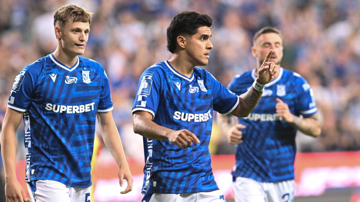 Trzech piłkarzy w niebieskich koszulkach Lech Poznań z napisem SUPERBET, jeden z nich wskazuje palcem przed siebie.