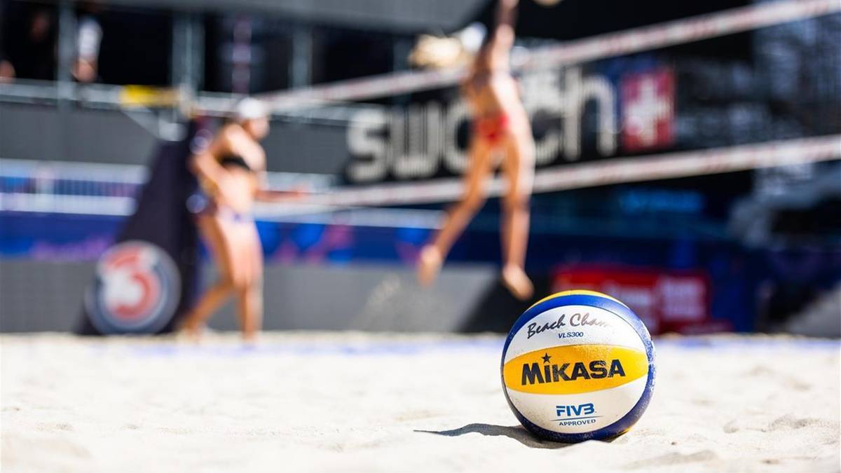 Piłka do siatk&oacute;wki plażowej leżąca na piasku, z niewyraźnymi sylwetkami zawodnik&oacute;w grających w tle.