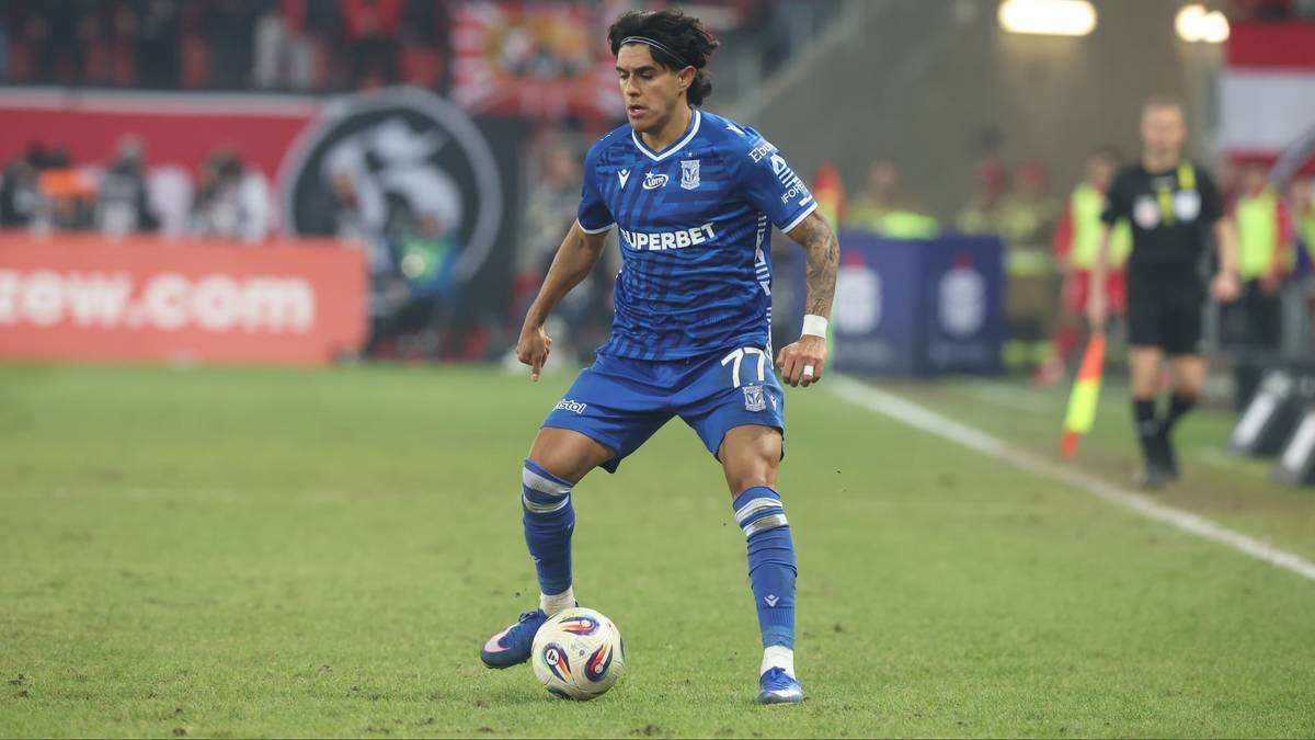 Piłkarz Lecha Poznań w niebieskim stroju z numerem 77 na plecach prowadzi piłkę na boisku.