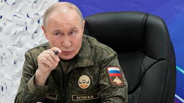 Władimir Putin w mundurze wojskowym siedzi w fotelu, wskazując palcem.