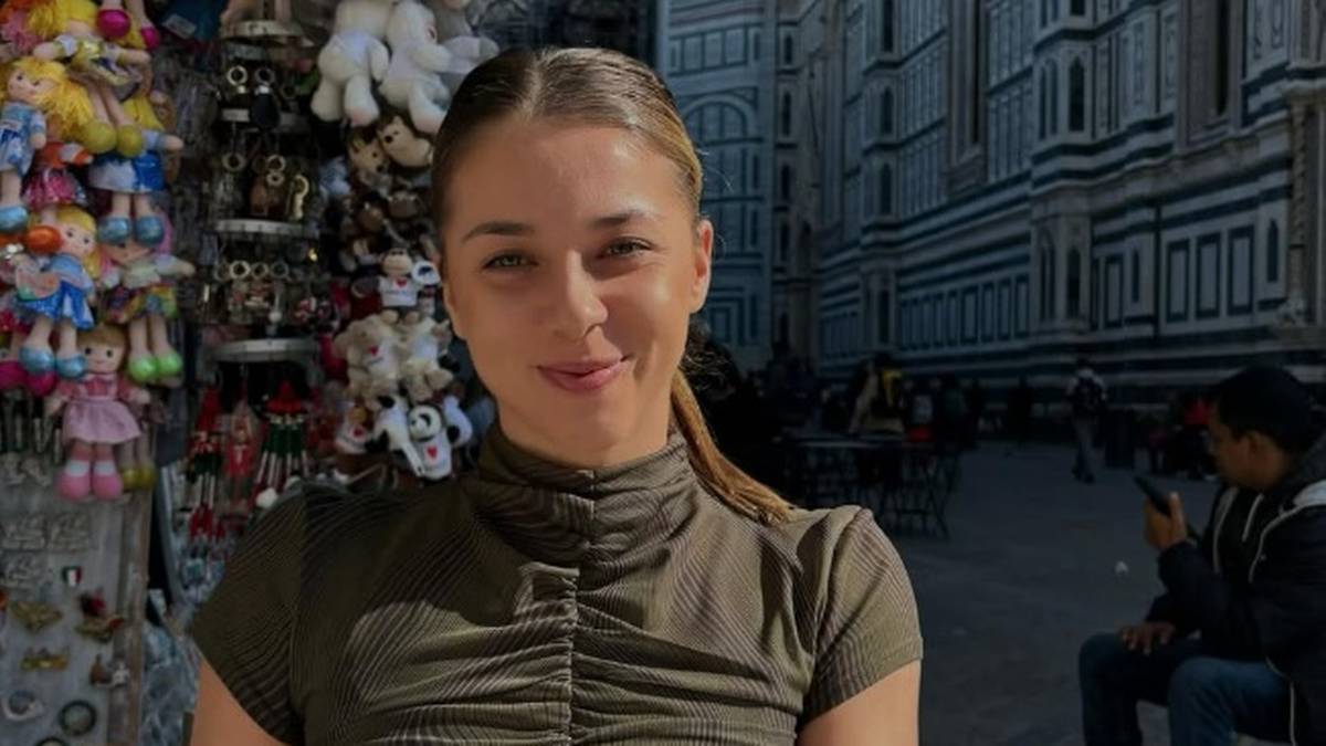 Młoda kobieta z uśmiechem na twarzy, w tle widoczne są budynki miejskie i stoisko z pamiątkami.