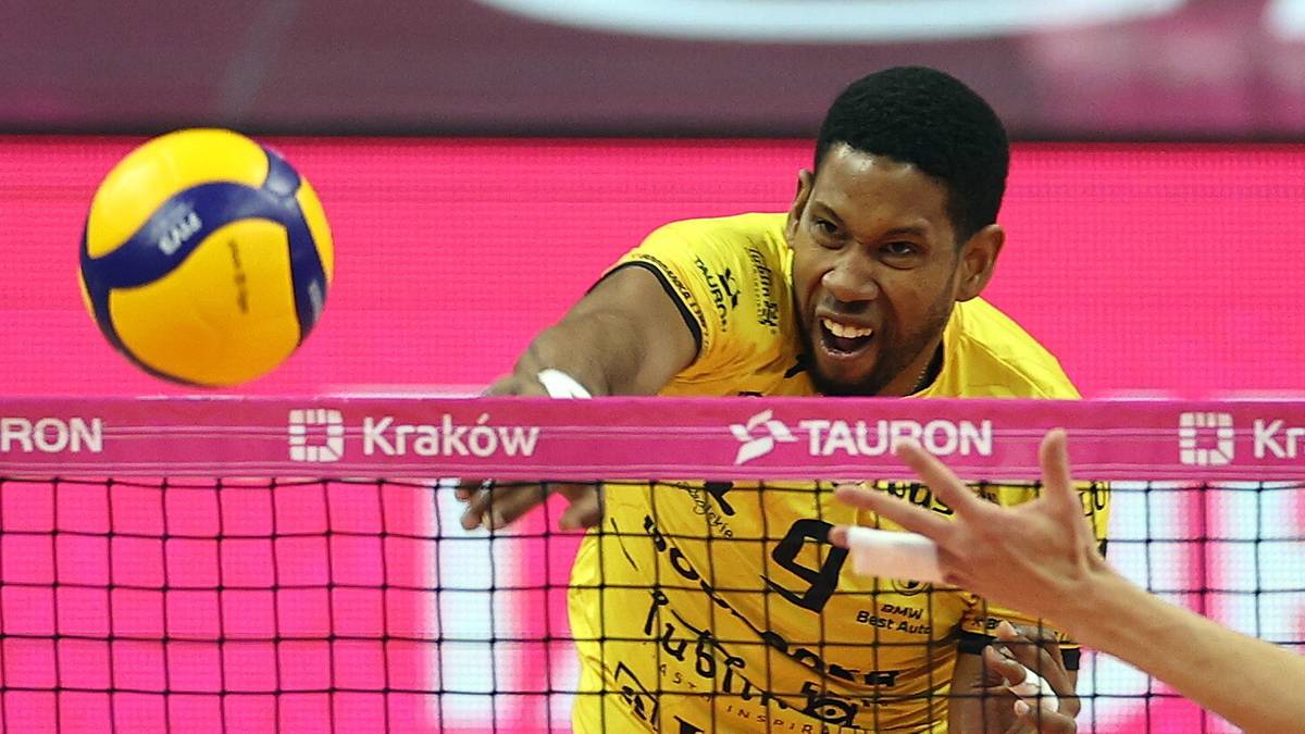 Siatkarz w ż&oacute;łtej koszulce wykonuje atak nad siatką, blokowany przez zawodnika w zielonej koszulce.