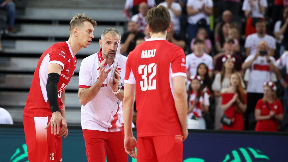 Trzech polskich siatkarzy w czerwonych strojach podczas meczu, jeden z nich otrzymuje instrukcje od trenera.