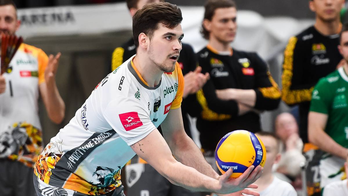 Siatkarz w białej koszulce z żółtymi i czarnymi elementami, z uwagą trzyma piłkę do siatkówki przed sobą, przygotowując się do zagrania. W tle widoczni są inni zawodnicy w podobnych strojach.