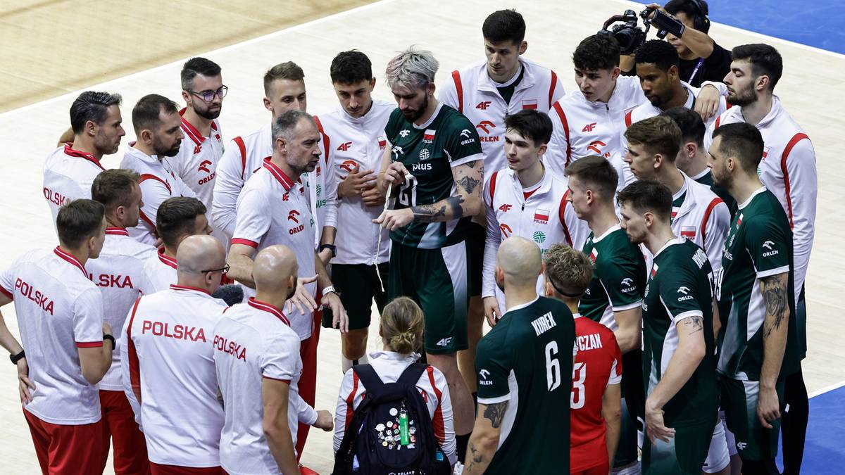 Grupa męskiej drużyny siatkarskiej w strojach sportowych, stojąca w kółku i słuchająca wskazówek trenera.