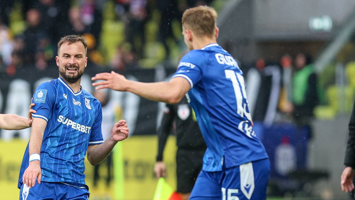 Dwóch piłkarzy w niebieskich koszulkach radośnie rozmawia na boisku piłkarskim.