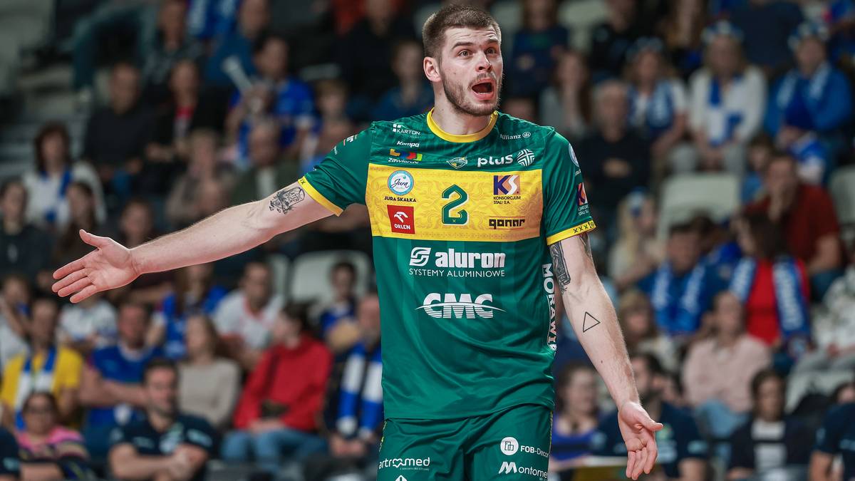 Siatkarz w zielono-żółtej koszulce z numerem 2, stojący na boisku, z otwartymi ramionami i ekspresyjną miną.