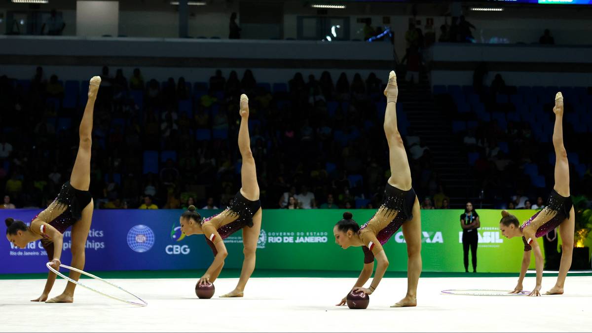 Pięć gimnastyczek artystycznych w czarnych strojach wykonuje w równym szeregu ćwiczenia z piłkami na dywanie.