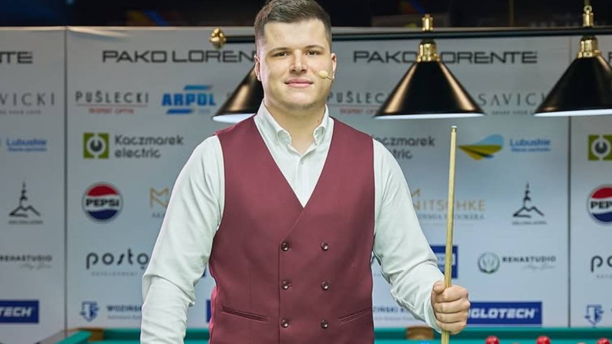 Polski zawodnik bilardowy w kamizelce i koszuli, trzymający kij bilardowy, stoi przy stole bilardowym.