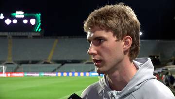 Mężczyzna z jasnymi włosami i niebieskimi oczami w szarej bluzie z kapturem patrzy w bok, z tyłu widać boisko piłkarskie i trybuny stadionu.