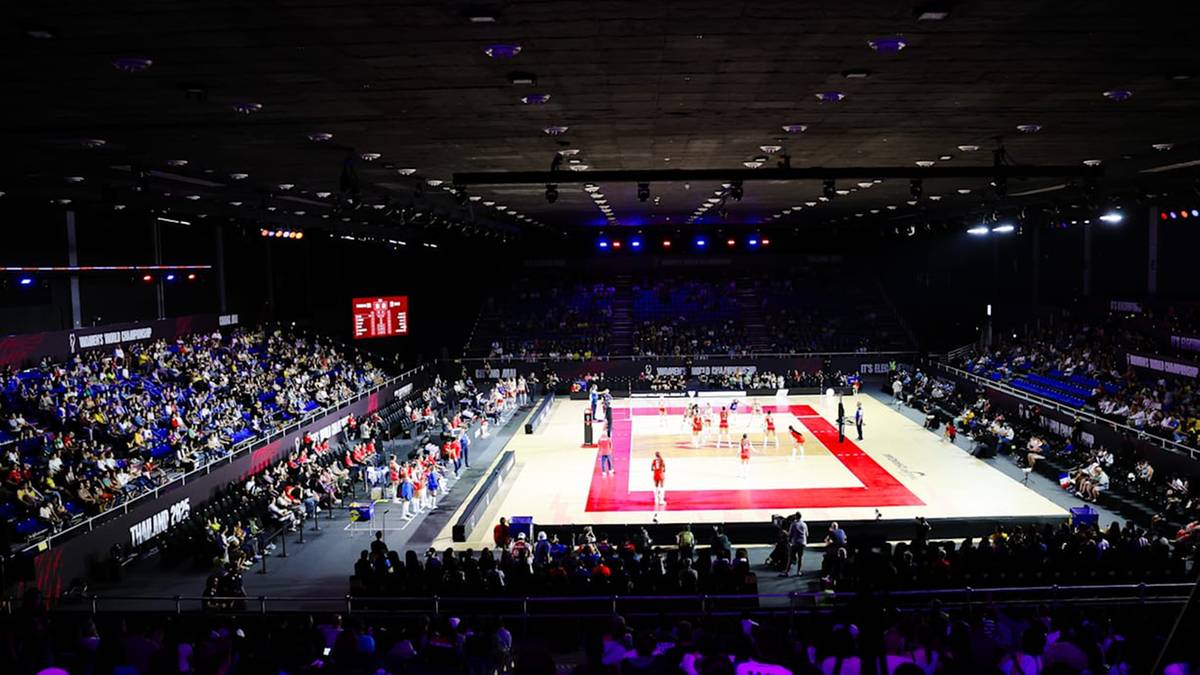 Widok na halę sportową podczas meczu siatk&oacute;wki z trybunami wypełnionymi widzami. Boisko do siatk&oacute;wki jest oświetlone, a na nim widać zawodnik&oacute;w.