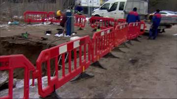 Teren po awarii miejskiej, zabezpieczony czerwonymi barierami, widoczne osoby w strojach roboczych.