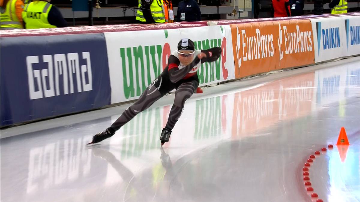 Łyżwiarka szybka w pozycji do jazdy na zakręcie, ubrana w czarno-czerwony kombinezon, na lodowisku z bandami reklamowymi w tle.