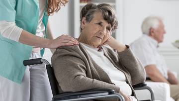 Starsza kobieta siedzi na w&oacute;zku inwalidzkim, z ręką na biodrze i zamyślonym wyrazem twarzy. Obok niej stoi inna osoba, kt&oacute;ra kładzie jej dłoń na ramieniu.