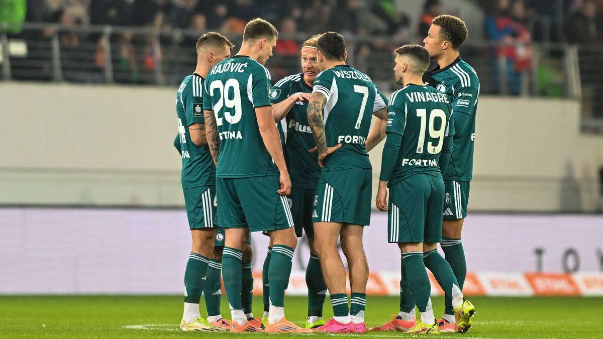 Drużyna piłkarska Legii Warszawa w zielonych strojach stoi w okręgu na boisku piłkarskim, rozmawiając ze sobą.