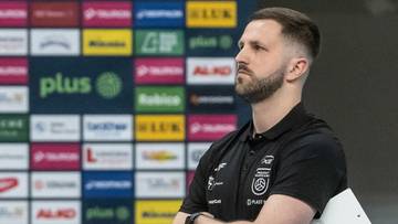 Trener siatk&oacute;wki w czarnej koszulce z założonymi rękami na piersi, stojący przed kolorową ścianą z logotypami sponsor&oacute;w.