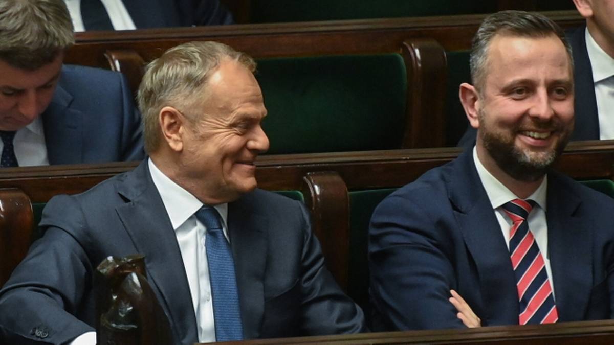 Donald Tusk i inny mężczyzna siedzą obok siebie w sali sejmowej, obaj uśmiechnięci.