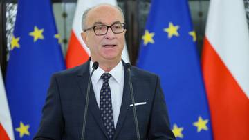 Mężczyzna w garniturze za m&oacute;wnicą, przemawia do mikrofon&oacute;w, w tle flagi Polski i Unii Europejskiej.