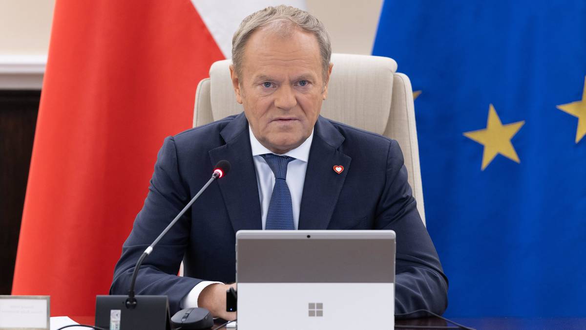 Premier Donald Tusk siedzi przy biurku z laptopem, w tle flagi Polski i Unii Europejskiej.