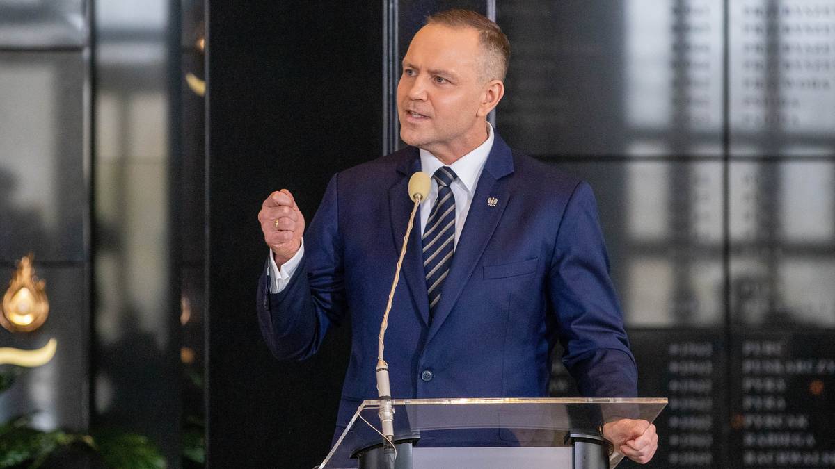 Mężczyzna w garniturze z krawatem przemawia przy mównicy, trzymając rękę zaciśniętą w pięść.