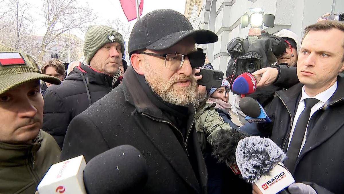 Grzegorz Braun w czapce i okularach przemawia do grupy dziennikarzy trzymających mikrofony.