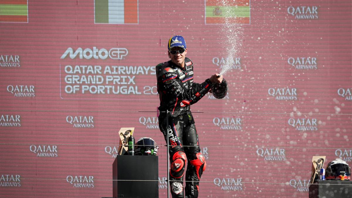 Motocyklista w czarno-czerwonym kombinezonie i niebieskim kasku na podium, trzymający butelkę szampana i rozpylający go.