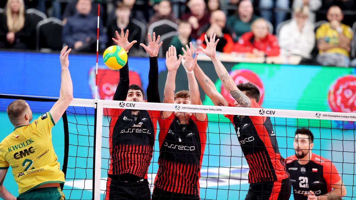 Siatkarze podczas meczu, jeden z nich wybija piłkę nad siatką, inni pr&oacute;bują ją zablokować.