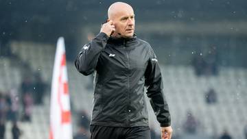 Sędzia piłkarski Szymon Marciniak w czarnej kurtce stoi na boisku, przykładając rękę do ucha.