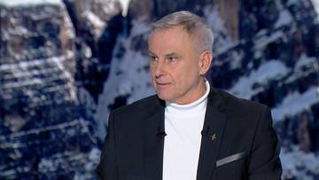 Mężczyzna w czarnym garniturze i białym golfie, na tle fotografii gór.