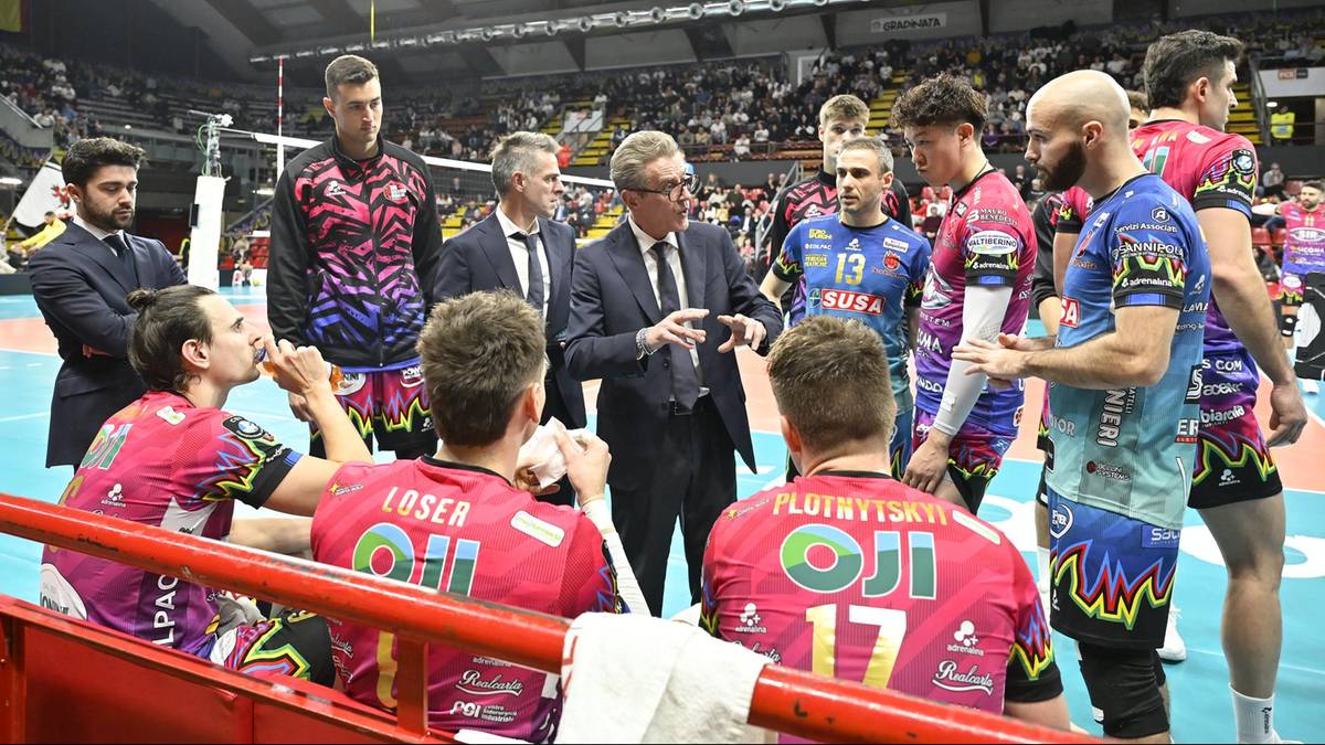 Drużyna siatkarska w przerwie meczu, zawodnicy w różowych strojach siedzą na ławce, trener rozmawia z nimi.