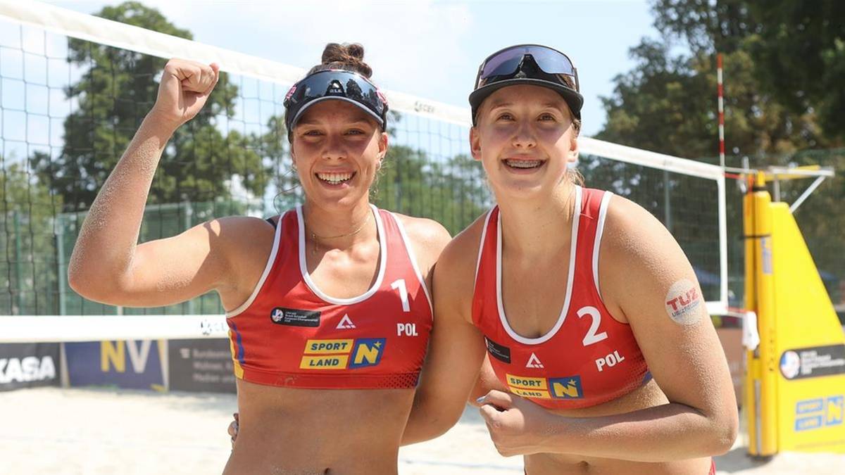 Dwie młode polskie siatkarki plażowe w czerwonych strojach sportowych, z numerami 1 i 2 na koszulkach, uśmiechają się do aparatu, unosząc ramiona w geście radości.