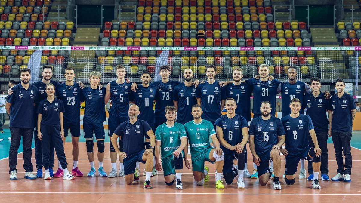 Drużyna siatkarska w komplecie strojów sportowych stoi na boisku siatkarskim przed trybunami wypełnionymi widzami.