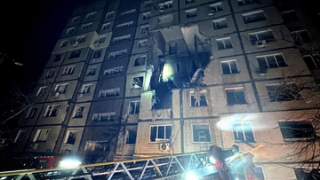 Wielopiętrowy budynek mieszkalny z widocznymi uszkodzeniami na wyższych piętrach po ataku, obok drabina strażacka