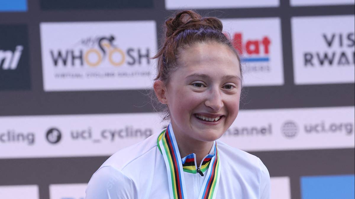 Młoda kobieta z medalem na szyi uśmiecha się szeroko, z włosami upiętymi w kok. W tle widoczne logo UCI Cycling.