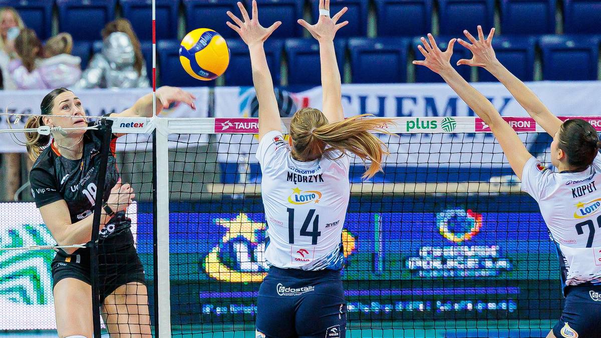 Siatkarka zagrywa piłkę, dwie inne próbują ją zablokować.