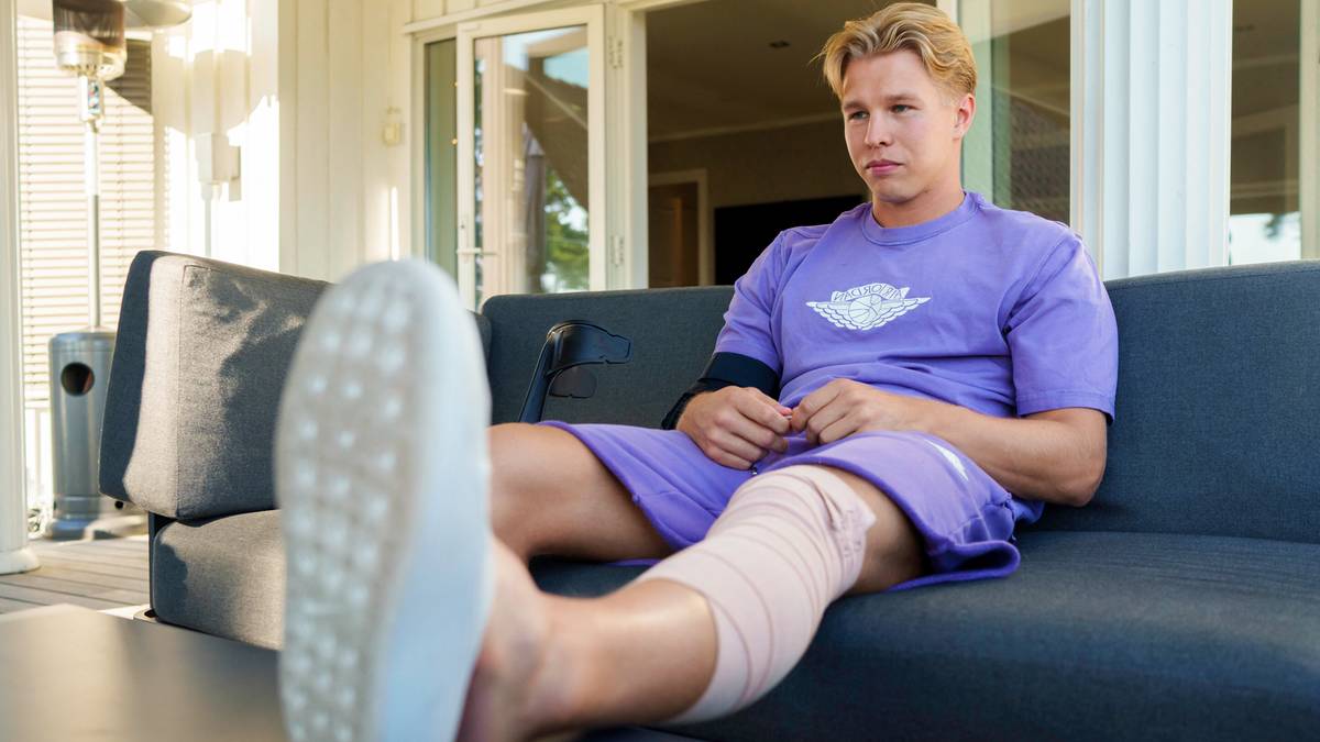 Młody mężczyzna siedzi na kanapie, z bandażem na kolanie, podczas rekonwalescencji.