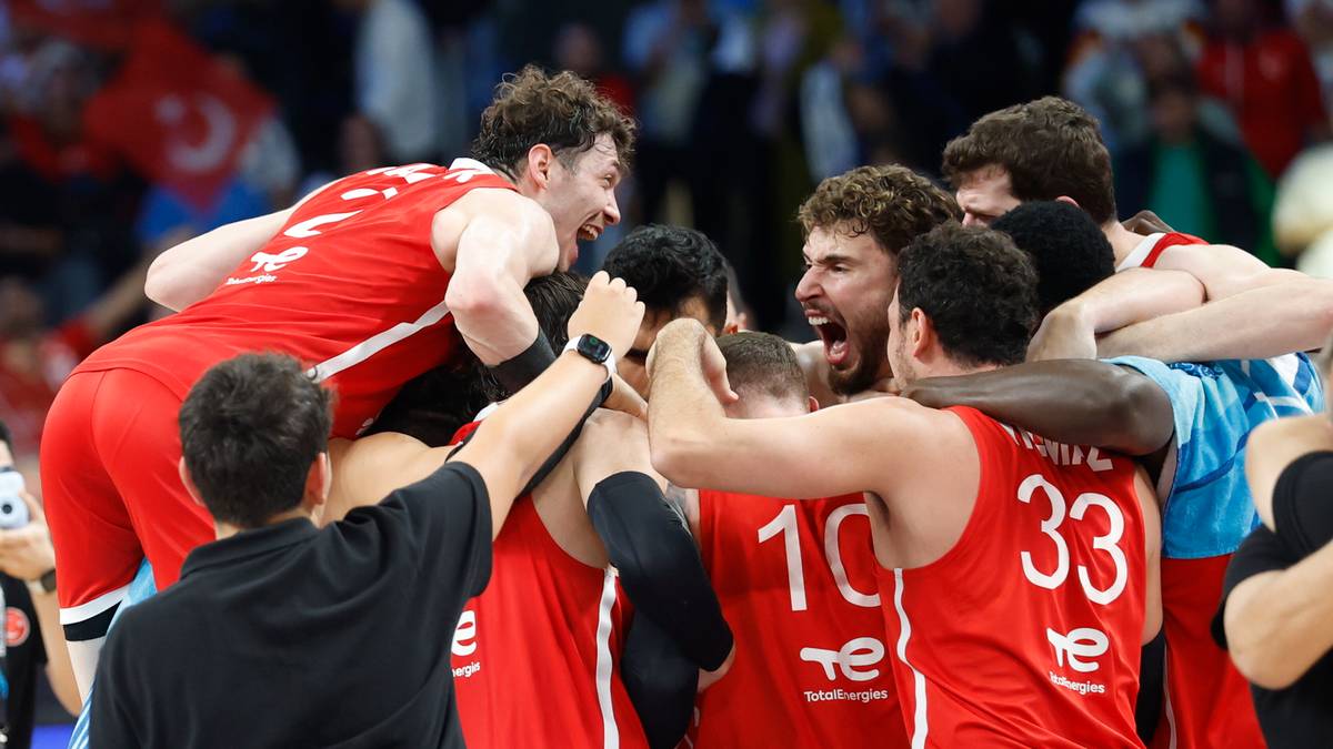 Drużyna koszykówki w czerwonych strojach celebruje zwycięstwo, podnosząc jednego z graczy na ramiona. Widoczni są zawodnicy z numerami 10 i 33 na koszulkach.